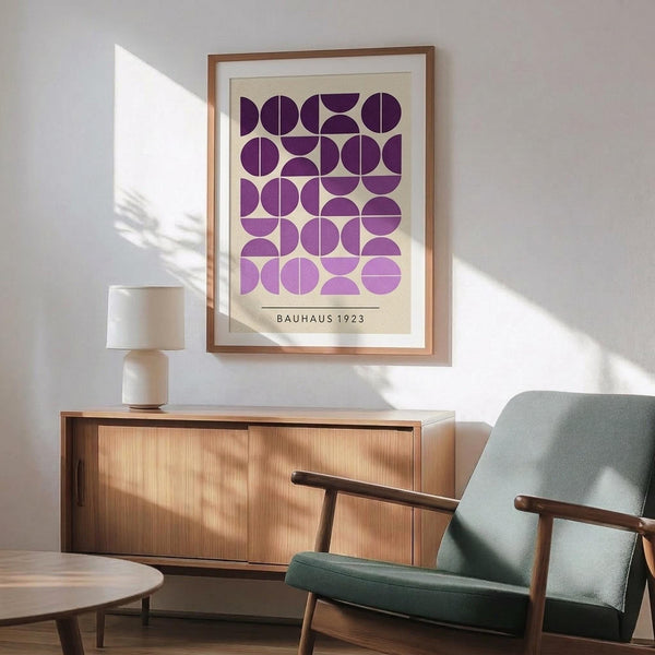 May include: Framed geometric poster with purple and lavender circles and semicircles arranged in a grid pattern on cream background, displayed above mid-century modern wood credenza with ceramic table lamp, next to sage green upholstered armchair with wooden arms in sunlit room with white walls.