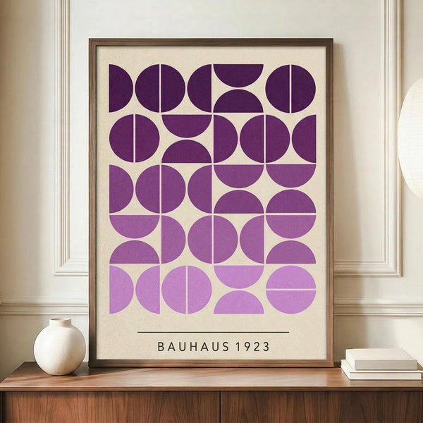 May include: Framed geometric poster with purple circles and semicircles in gradient shades from deep purple to lavender on cream background, text reading "BAUHAUS 1923" below the pattern. Brown wooden frame sits on walnut credenza next to small white ceramic vase and stacked books in bright room with white paneled walls.