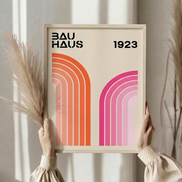 May include: Hands holding a framed geometric poster with "BAUHAUS 1923" text and two gradient arches - one orange-red on left, one magenta-pink on right, displayed against neutral curtains with dried pampas grass visible on left side