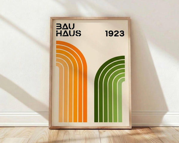 May include: Framed poster with cream background featuring geometric design of curved parallel lines in orange gradient and green gradient forming arch shapes. Black text reads "BAUHAUS" and "1923" at top. Natural wood frame sits on light wooden floor against white wall with soft natural lighting creating diagonal shadows.