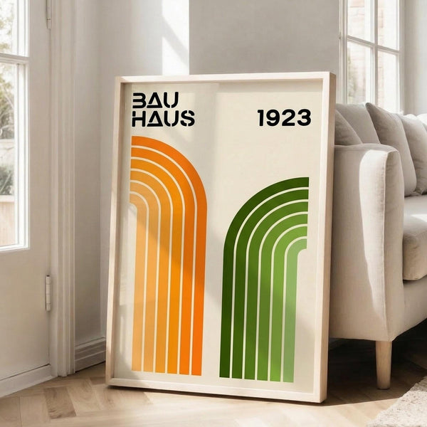 May include: Framed poster with geometric design featuring orange and green concentric arches on cream background, displayed in bright living room corner next to beige sofa. Natural light streams through white-framed windows onto light wood floors. The minimalist artwork shows "BAUHAUS 1923" text in black modern typography above two sets of curved parallel lines in gradient orange-to-yellow and dark-to-light green colors.