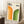 May include: Framed poster with cream background featuring orange and green geometric arches, black "BAUHAUS 1923" text at top. Modern minimalist design with concentric curved lines in gradient orange-to-yellow and dark-to-light green. Wooden frame leaning against white wall next to beige couch, white coffee cup on wooden side table, cream textured rug below.