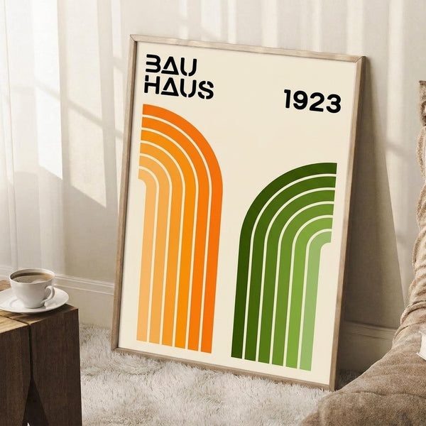 May include: Framed poster with cream background featuring orange and green geometric arches, black "BAUHAUS 1923" text at top. Modern minimalist design with concentric curved lines in gradient orange-to-yellow and dark-to-light green. Wooden frame leaning against white wall next to beige couch, white coffee cup on wooden side table, cream textured rug below.