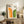 May include: Framed poster with bold geometric design featuring orange and green rainbow arches on cream background, displayed on rustic wooden sideboard alongside fern plant, books, ceramic vase, and leather armchair in modern living room setting with neutral walls