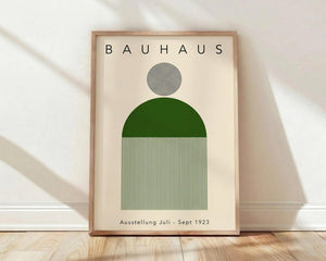 May include: Framed Bauhaus-style poster with cream background featuring black concentric circles at top, green semicircle in middle, and black and white vertical stripes below. Text reads "BAUHAUS" and "Ausstellung Juli - Sept 1923". Light wood frame sits on wooden floor against white wall with soft shadows.