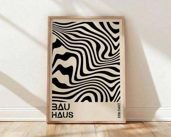 May include: Framed poster with black and white swirling optical illusion pattern featuring flowing wavy lines that create a hypnotic distorted effect. Text reads "BAUHAUS" and "EXPO 1923" in modern sans-serif font. Light wooden frame sits on natural wood flooring against white wall with soft shadows from window light.