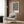 May include: Black and white abstract wavy lines poster in light wood frame hanging on wood paneled wall above beige upholstered armchair with geometric patterned throw pillow. Modern brass floor lamp and dark round side table with open book nearby. Neutral toned contemporary living room setting with natural lighting from window.
