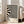 May include: Black and white wavy abstract pattern poster in light wood frame leaning against white wall next to beige sofa. The design features flowing curved lines creating an optical illusion effect with "BAUHAUS" text at bottom. Natural light streams through windows in minimalist living room setting with light wooden floors.