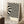 May include: Black and white abstract wavy lines poster in light wood frame leaning against white wall corner. Bauhaus text at bottom. Coffee cup on wooden block nearby, beige throw blanket visible. Cream colored background with flowing distorted stripes creating optical illusion effect.