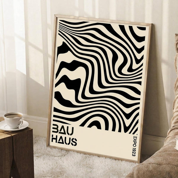 May include: Black and white abstract wavy lines poster in light wood frame leaning against white wall corner. Bauhaus text at bottom. Coffee cup on wooden block nearby, beige throw blanket visible. Cream colored background with flowing distorted stripes creating optical illusion effect.