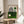 May include: Framed Bauhaus exhibition poster with geometric design featuring black concentric lines forming an arch, green and cream color blocks, and small circles, displayed on round wooden side table next to beige upholstered chair with plant shadows cast across white wall in bright natural lighting.