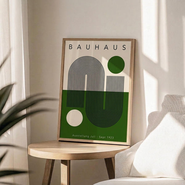 May include: Framed Bauhaus exhibition poster with geometric design featuring black concentric lines forming an arch, green and cream color blocks, and small circles, displayed on round wooden side table next to beige upholstered chair with plant shadows cast across white wall in bright natural lighting.