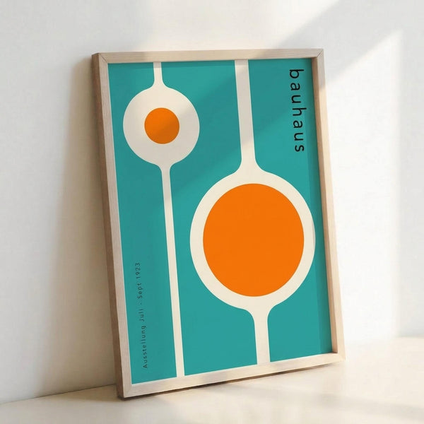 Framed poster with turquoise background featuring two white circular shapes with orange centers resembling stylized flowers or abstract forms. The design includes "bauhaus" text vertically along the right edge and "Ausstellung Juli Sept 1923" along the left side. The artwork has a mid-century modern aesthetic with clean geometric lines. Natural wood frame sits against white wall casting subtle shadow.