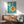 May include: Framed mid-century modern poster with turquoise background featuring white geometric shapes and orange circles resembling kitchen utensils, displayed above wooden sideboard with white ceramic lamp in minimalist living room with sage green upholstered chair and natural lighting creating diagonal shadows on white wall