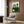 May include: Bauhaus-style poster with green and cream geometric design featuring concentric arcs and circles, displayed in light wood frame on wooden wall panel. Modern beige armchair with geometric pillow, brass floor lamp with white base, and dark round side table with open book create contemporary living room setting with natural lighting from window.