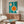May include: Teal poster with white and orange geometric circles resembling kitchen utensils displayed above wooden mid-century credenza in modern living room. White curved chair and glass-top coffee table visible in foreground. Natural lighting creates shadows on light walls.