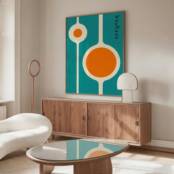 May include: Teal poster with white and orange geometric circles resembling kitchen utensils displayed above wooden mid-century credenza in modern living room. White curved chair and glass-top coffee table visible in foreground. Natural lighting creates shadows on light walls.