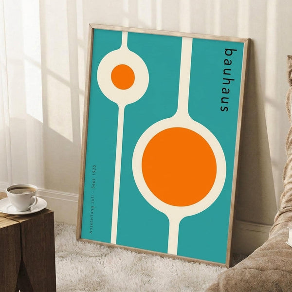 May include: Framed mid-century modern poster with turquoise background featuring white organic shapes and bright orange circles in Bauhaus style, leaning against white wall corner next to wooden block with coffee cup and saucer, positioned on light textured rug beside beige fabric furniture