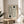 May include: Framed geometric print with concentric oval line patterns and dark green circles on cream background, displayed on round wooden stool beside beige upholstered chair with knit throw pillow. Natural lighting creates soft shadows on neutral wall with plant leaves visible in corner of minimalist room setting.