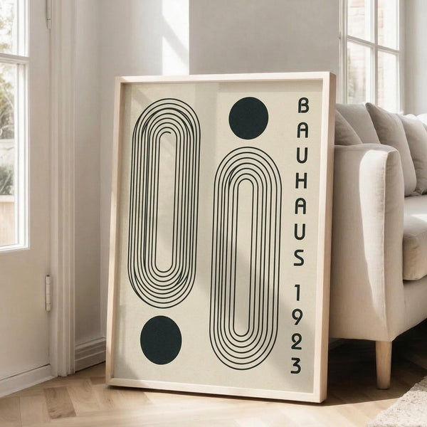 May include: Large framed art print leaning against white wall in bright living room with beige sofa. Print features bold black concentric oval shapes and dark green circles on cream background, with vertical "BAUHAUS 1923" text. Light wood frame, approximately 3 feet tall, positioned on light wooden floors near tall windows with natural lighting.