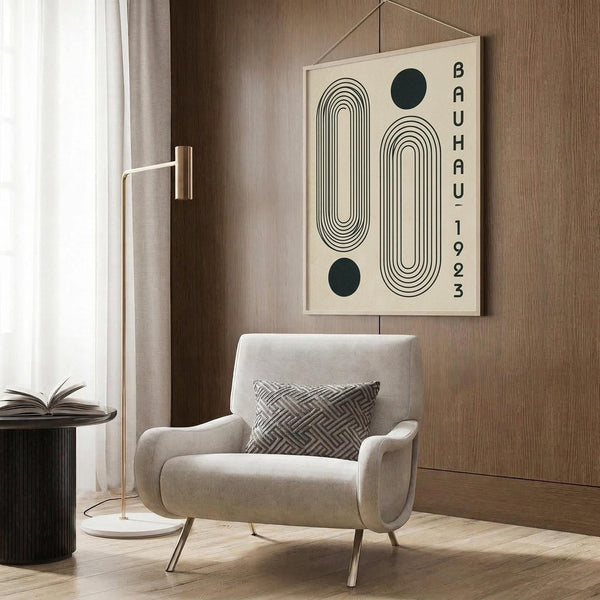 May include: Cream-colored poster with black geometric design featuring two rounded rectangular shapes with concentric lines and two dark circles, text reading "BAUHAUS 1923" vertically aligned on right side, hanging on wood-paneled wall above beige upholstered armchair with patterned pillow, brass floor lamp and side table with open book visible in modern neutral-toned room setting