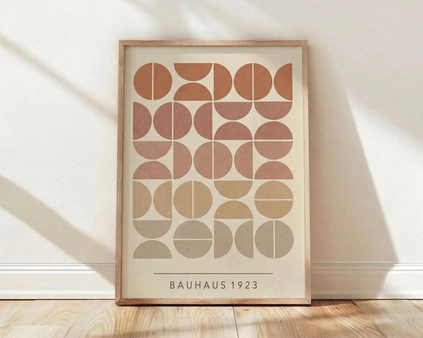 May include: Framed geometric poster with circles and semicircles in earth tones of terracotta, rust, tan, and beige arranged in a grid pattern. Text reads "BAUHAUS 1923" at bottom. Light wood frame sits on wooden floor against white wall with soft natural lighting casting shadows.