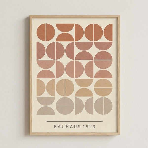 May include: Framed geometric art print with circular and semicircular shapes arranged in a grid pattern. Features warm earth tones transitioning from terracotta and rust at the top through peachy pink in the middle to beige and taupe at the bottom. Text reads "BAUHAUS 1923" below the design. Displayed in light wood frame against cream colored wall.