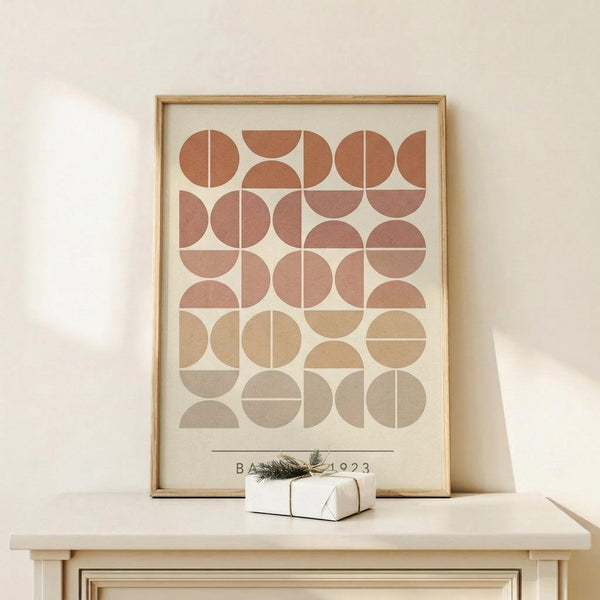 May include: Framed geometric art print featuring circles and semicircles in warm terracotta, rust, beige and taupe tones arranged in a grid pattern, displayed on light wood surface alongside small white wrapped gift with twine and greenery sprig against cream wall
