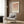 May include: Framed geometric art print with terracotta, beige and cream semicircles and circles arranged in a grid pattern, displayed on wood paneled wall above modern beige armchair with patterned pillow. Scene includes brass floor lamp, dark round side table with open book, and neutral curtains in contemporary living room setting.