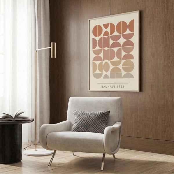 May include: Framed geometric art print with terracotta, beige and cream semicircles and circles arranged in a grid pattern, displayed on wood paneled wall above modern beige armchair with patterned pillow. Scene includes brass floor lamp, dark round side table with open book, and neutral curtains in contemporary living room setting.