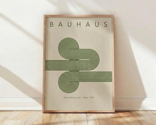 May include: Framed Bauhaus-style poster with green geometric design featuring flowing curved lines made of parallel stripes forming an abstract shape. Cream colored background with "BAUHAUS" text at top and "Ausstellung Juli - Sept 1923" at bottom. Natural wood frame leaning against white wall on light wooden floor with soft natural lighting casting shadows.
