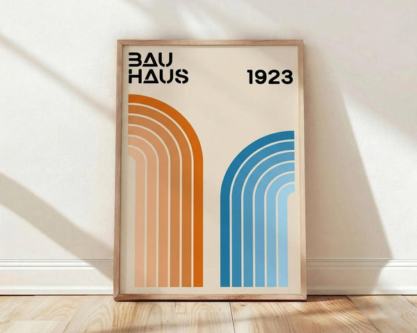 May include: Framed geometric poster with cream background displaying "BAUHAUS 1923" text and two curved rainbow stripe designs - orange gradient arcs on left, blue gradient arcs on right. Light wood frame sits on wooden floor against white wall with soft natural lighting casting diagonal shadows.