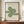 May include: Framed Bauhaus poster with green geometric spiral design made of concentric curved lines on cream background, displayed on wooden sideboard next to white ceramic vase and stack of books against white paneled wall