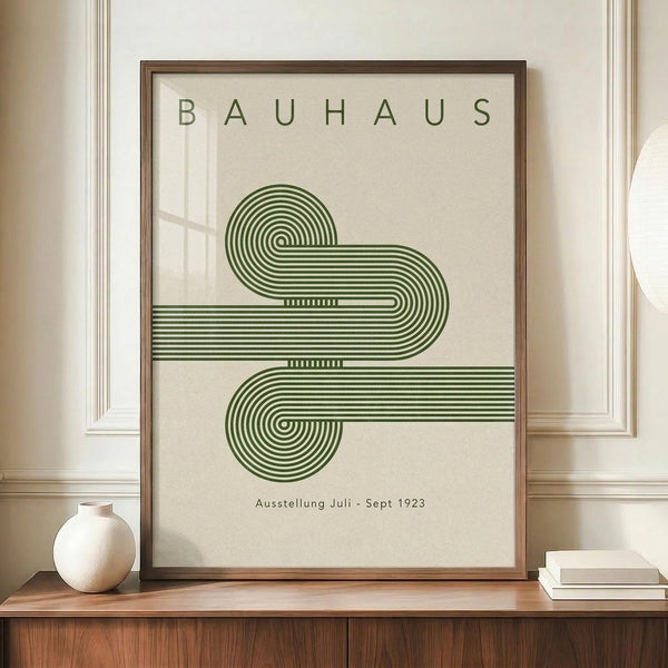 May include: Framed Bauhaus poster with green geometric spiral design made of concentric curved lines on cream background, displayed on wooden sideboard next to white ceramic vase and stack of books against white paneled wall