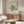 May include: Modern minimalist room interior featuring a framed Bauhaus exhibition poster with green geometric spiral design on cream background, positioned above a mid-century walnut sideboard. White sculptural chair and glass-top coffee table complete the scene, with natural light creating soft shadows on neutral walls.