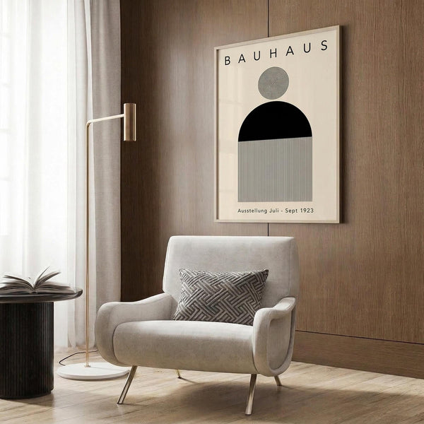 May include: Minimalist Bauhaus poster with geometric shapes in black, white and gray hanging on wood-paneled wall above cream upholstered armchair with patterned pillow. Modern reading nook features brass floor lamp, dark round side table with open book, and warm neutral tones throughout the styled interior scene.