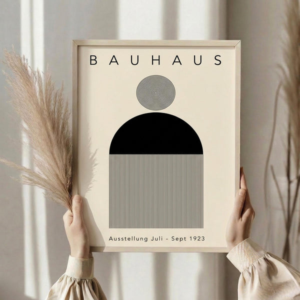 May include: Person holding framed Bauhaus poster with geometric design featuring concentric circles, black semicircle, and vertical black stripes on cream background. Light wooden frame, neutral beige clothing, dried pampas grass, soft natural lighting against white curtained window.