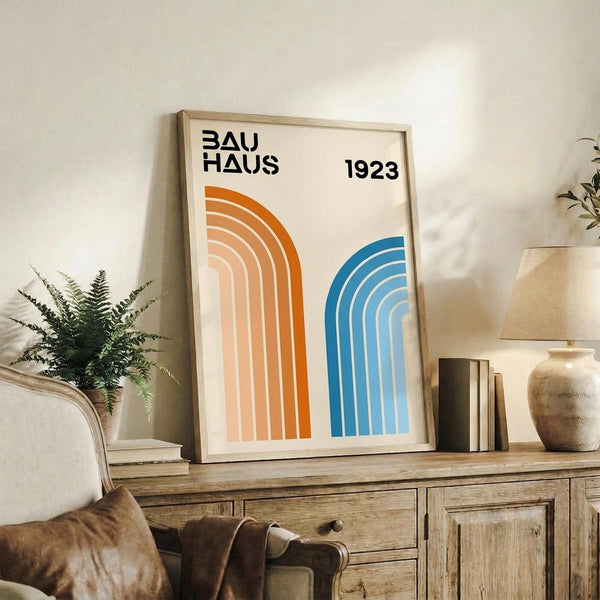 May include: Framed Bauhaus poster with orange and blue geometric arches on cream background, displayed on rustic wooden sideboard alongside fern plant, stacked books, ceramic vase, and leather chair in neutral-toned living room setting