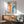 May include: Framed Bauhaus poster with orange and blue curved geometric stripes on cream background, displayed above mid-century modern wooden sideboard with ceramic lamp. Sage green upholstered chair with wooden arms sits nearby on wooden floor. Natural sunlight creates shadows across white wall in minimalist interior setting.