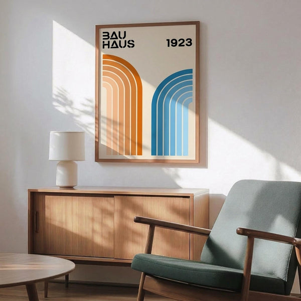 May include: Framed Bauhaus poster with orange and blue curved geometric stripes on cream background, displayed above mid-century modern wooden sideboard with ceramic lamp. Sage green upholstered chair with wooden arms sits nearby on wooden floor. Natural sunlight creates shadows across white wall in minimalist interior setting.