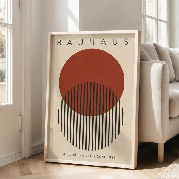 May include: Framed Bauhaus-style poster print featuring a rust-colored circle overlapping black and white vertical stripes on cream background, displayed in light wood frame leaning against white wall corner next to beige upholstered sofa in bright, naturally-lit room with hardwood floors and white area rug.