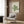 May include: A cream-colored poster with green geometric line art forming an abstract swirling design and "BAUHAUS" text at top, displayed in a light wooden frame on a textured wood wall. Modern beige armchair with geometric pillow sits below, alongside a gold floor lamp and dark round side table with open book.