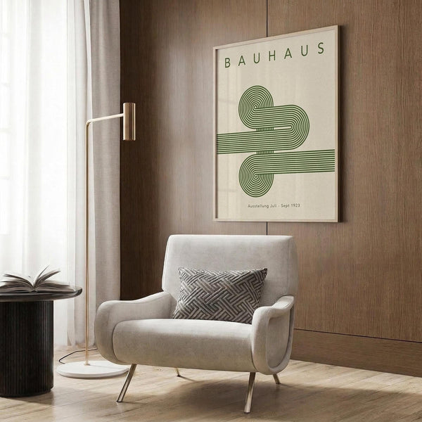May include: A cream-colored poster with green geometric line art forming an abstract swirling design and "BAUHAUS" text at top, displayed in a light wooden frame on a textured wood wall. Modern beige armchair with geometric pillow sits below, alongside a gold floor lamp and dark round side table with open book.