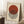 May include: Framed Bauhaus-style poster with cream background featuring a rust-colored circle overlapping black vertical stripes, German text at top and bottom. Wood frame leaning against white wall corner on textured white rug, next to wooden side table with white coffee cup and saucer, beige throw blanket visible.