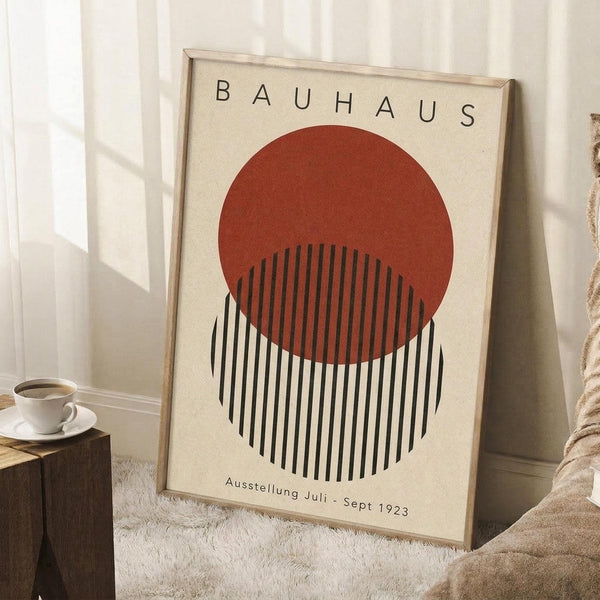 May include: Framed Bauhaus-style poster with cream background featuring a rust-colored circle overlapping black vertical stripes, German text at top and bottom. Wood frame leaning against white wall corner on textured white rug, next to wooden side table with white coffee cup and saucer, beige throw blanket visible.