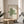 May include: Framed Bauhaus-style poster with green geometric spiral design displayed on round wooden side table next to gray upholstered chair. Cream-colored wall background with natural window light and plant shadows. Modern minimalist interior styling with warm wood frame and neutral tones.