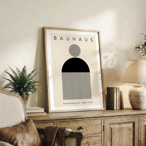 May include: Framed Bauhaus-inspired minimalist poster with geometric shapes - small circle with concentric lines, black semicircle, and striped rectangle on cream background. Displayed on rustic wooden sideboard with fern plant, books, ceramic vase, and leather sofa in neutral-toned living room setting.
