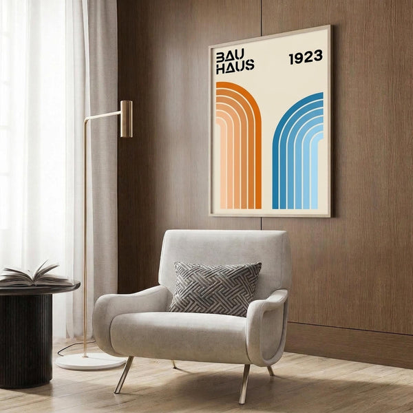 May include: A framed geometric poster on wood paneling featuring orange and blue curved stripes with "BAUHAUS 1923" text. Shows a modern living room setting with a beige upholstered chair with geometric patterned pillow, brass floor lamp, dark round side table with open book, and neutral curtains by a window.
