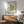 May include: Framed Bauhaus poster with green geometric spiral design hanging above wooden sideboard in modern living room. Mid-century teal armchair with wooden arms beside small round coffee table. White ceramic table lamp on credenza. Soft natural lighting creates shadows on white wall.