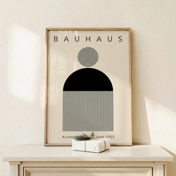 May include: Framed Bauhaus poster with geometric design featuring concentric circles, black semicircle, and vertical black and white stripes on cream background. Natural wood frame displayed on light wooden surface with small wrapped gift decorated with evergreen sprig against neutral wall.